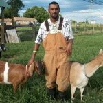 A man in overalls standing next to two goats.