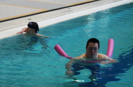 Two people swimming in an indoor pool with pink floats.