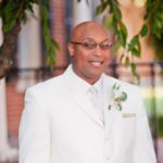 A man in a white suit is smiling in front of a tree.